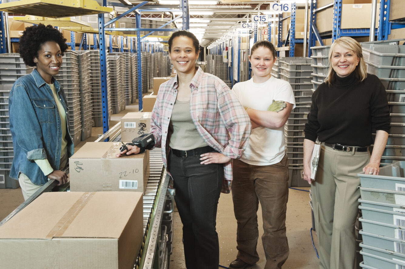Team portrait of multi-ethnic female warehouse workers working next to a motorzied feed conveyor in a large distribution warehouse.